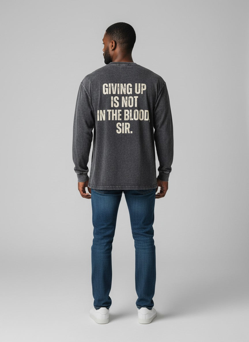A man viewed from the back wearing a dark gray long-sleeve shirt with cream text that reads 'GIVING UP IS NOT IN THE BLOOD SIR.' paired with blue jeans and white sneakers, against a light gray studio background.