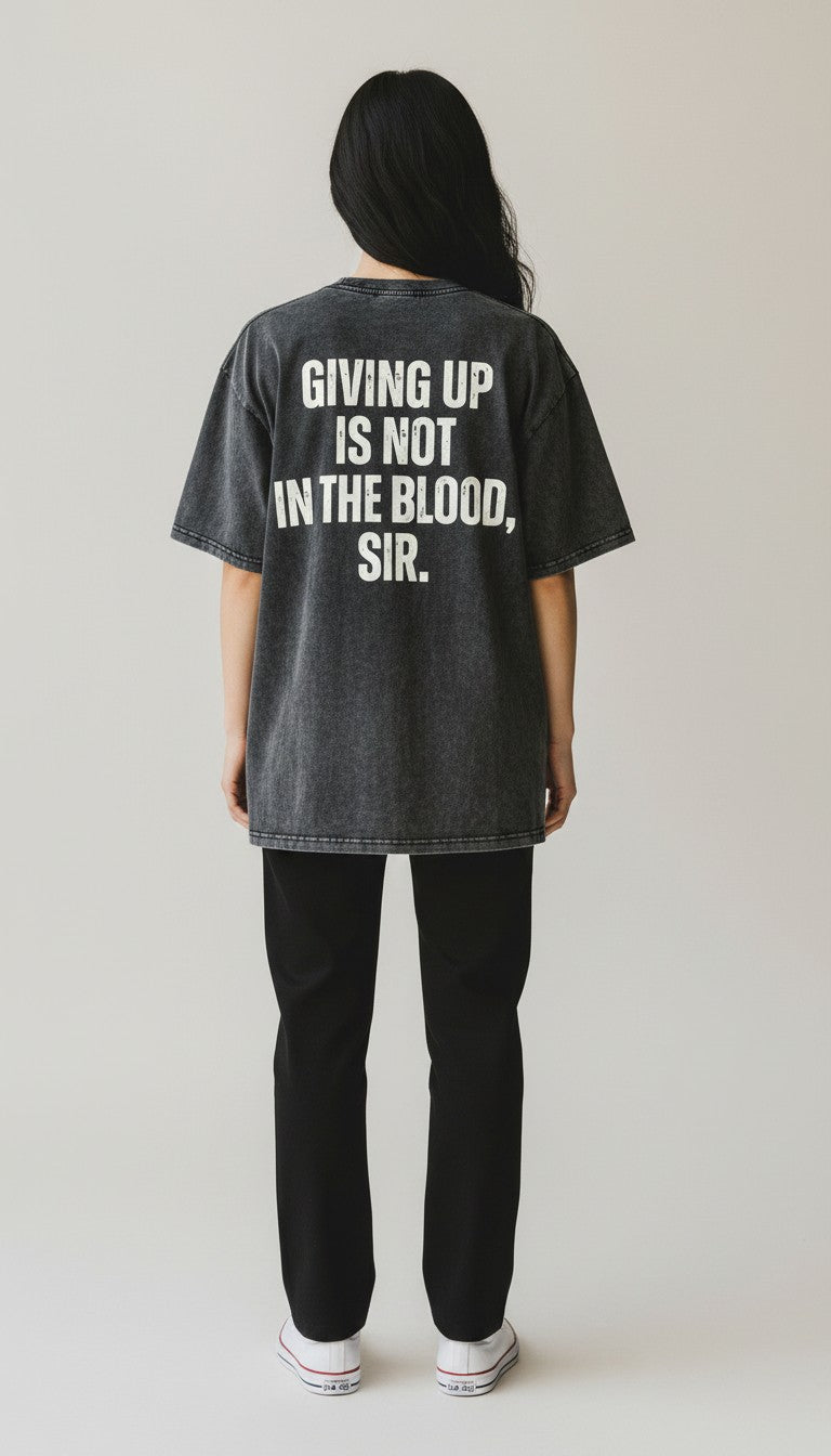 A person shown from the back wearing an oversized black vintage washed graphic T-shirt with bold white text reading 'GIVING UP IS NOT IN THE BLOOD, SIR.' paired with black pants and white sneakers, against a plain light gray studio background.