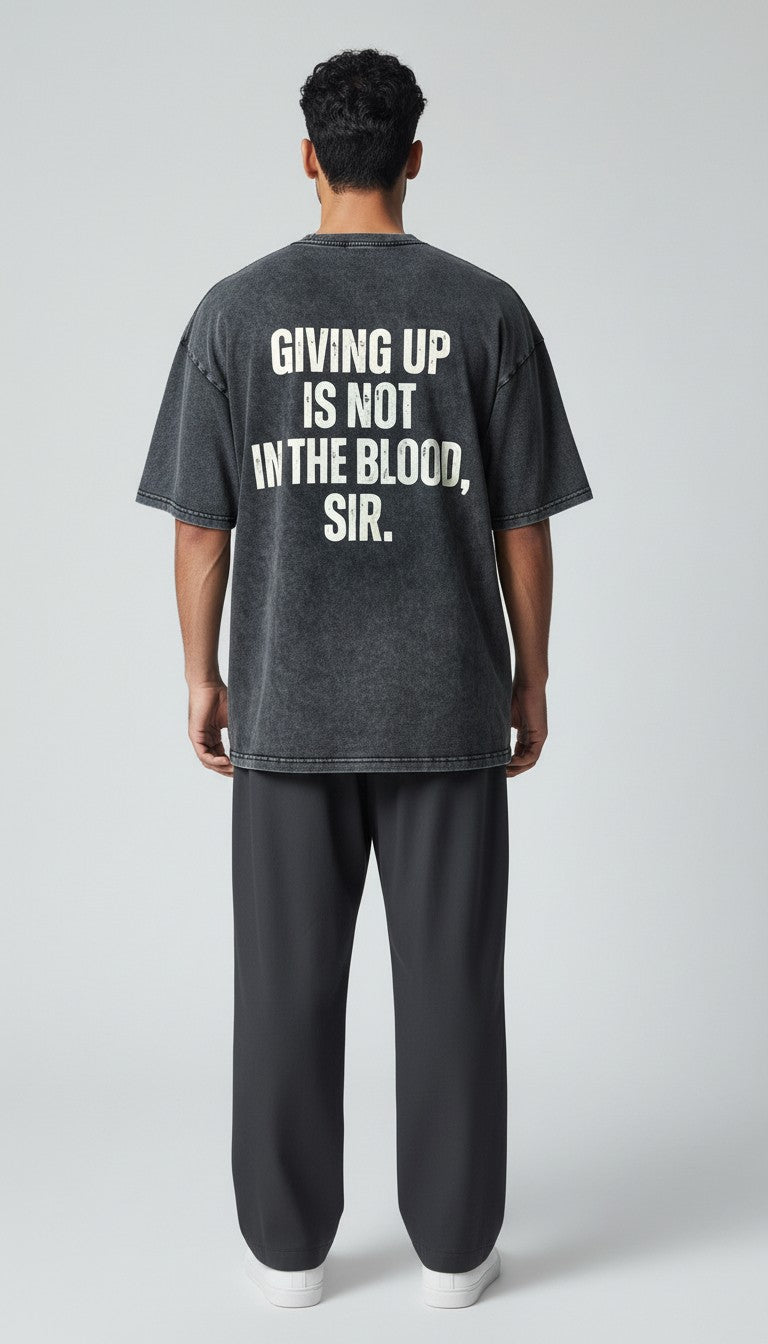 A man viewed from the back wearing a black vintage washed t-shirt with bold white text that reads 'GIVING UP IS NOT IN THE BLOOD, SIR.' and dark gray sweatpants, set against a light gray studio background.