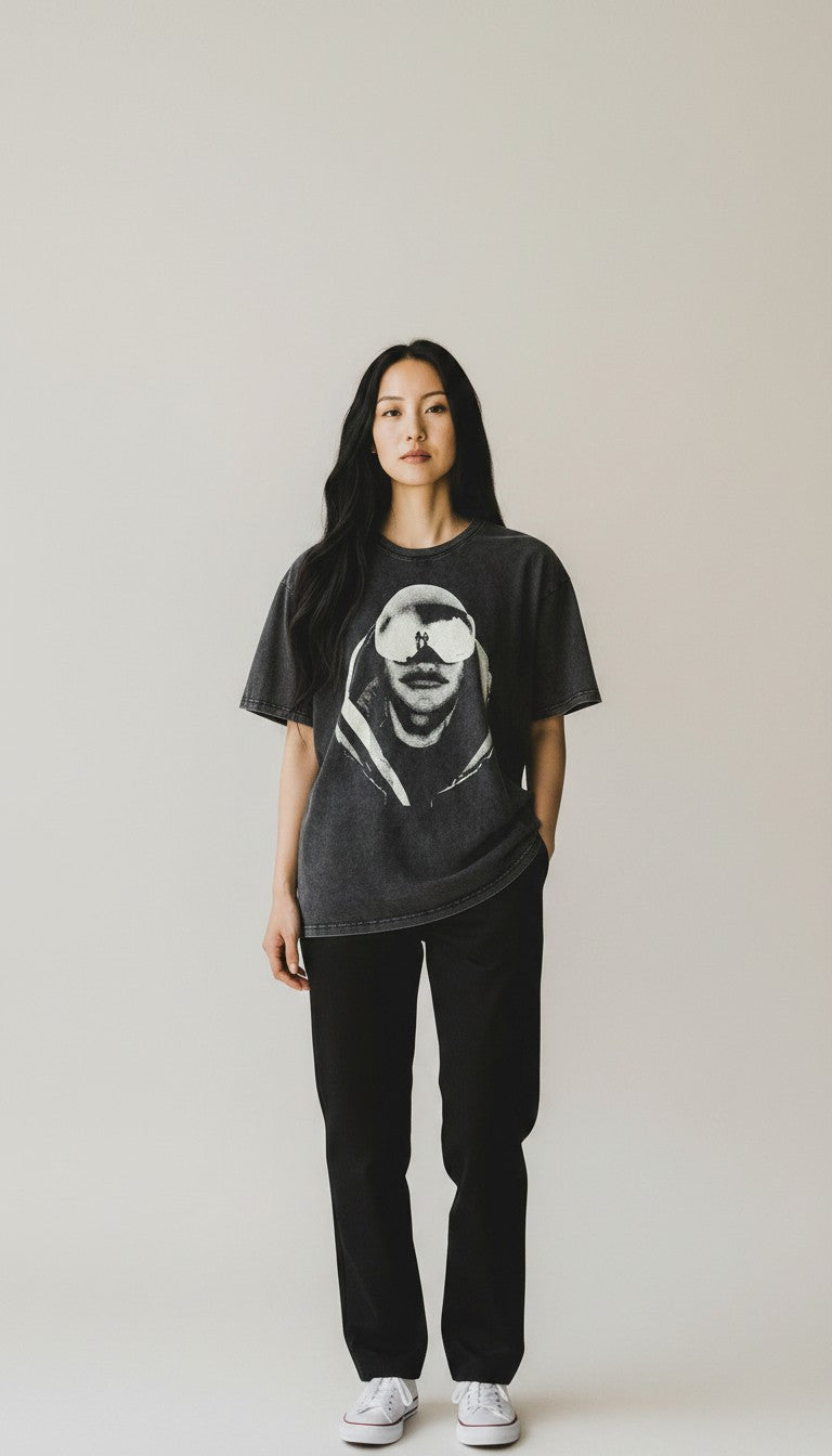 A woman stands against a plain light gray studio background wearing a faded black graphic T-shirt with a large white portrait print, black pants, and white sneakers.