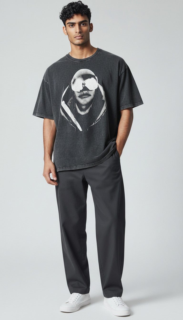 A man modeling a black vintage washed graphic t-shirt with a black and white portrait print, paired with dark gray sweatpants and white sneakers, against a light gray studio background.